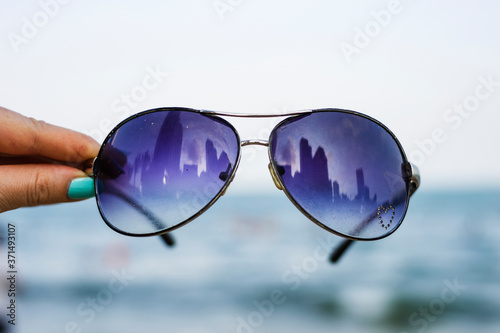 Female hand holds colorful sunglasses against the background of the sky and blue sea. The points reflect the storey buildings of the city