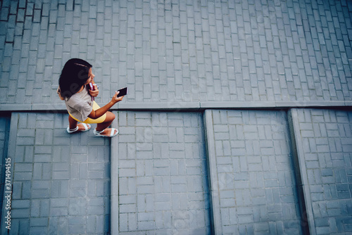 Top view of brunette hipster girl walking on stair carrying books and sending messages on mobile phone, female traveler having city tour  searching location on smartphone application browsing maps