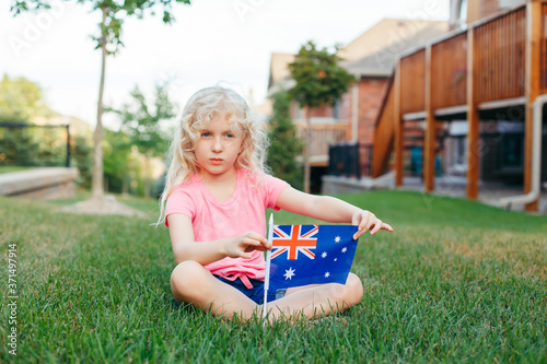 Adorable cute happy Caucasian girl holding Australian flag. Smiling child sitting on grass in park holding Australia flag. Kid citizen celebrating Australia Day holiday in January outdoors. © anoushkatoronto