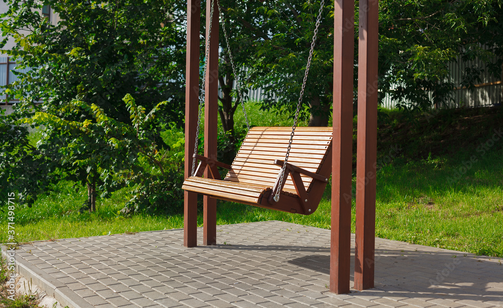 Wide wooden swing in the yard, swing for grown ups hanging on the chains, installation in country yard