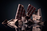 A broken chocolate bar and pieces of dark chocolate on a black reflective background.