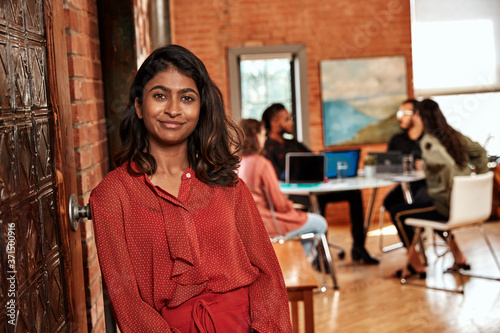 portrait young ethnic woman standing and leaning on sliding door, coworkers gathered around desk in background 