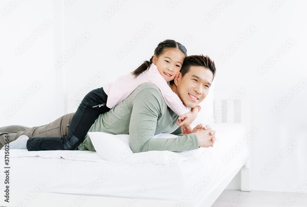 Portrait of lovely Asian daughter smiling and lying on her handsome father's back on white bed. Family holiday and togetherness. Happy Father's Day!