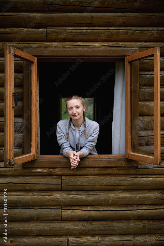 Young woman looks out of the window of a wooden house. Vacation outside the city. Country life. Happiness. A positive outlook on the world around you. Young woman looks out of the window of a wooden house. Vacation outside the city. Country life. Happiness. A positive outlook on the world around you.