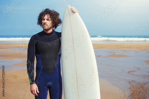 Portrait of professional young surfer standing against the ocean holding his beautiful surfboard with copy space area for text, cross process image, flare light