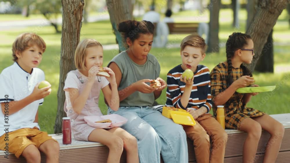Group of multiethnic boys and girls of elementary age sitting on bench in the park, eating sandwiches and fruit on lunch and chatting about something Group of multiethnic boys and girls of elementary age sitting on bench in the park, eating sandwiches and fruit on lunch and chatting about something