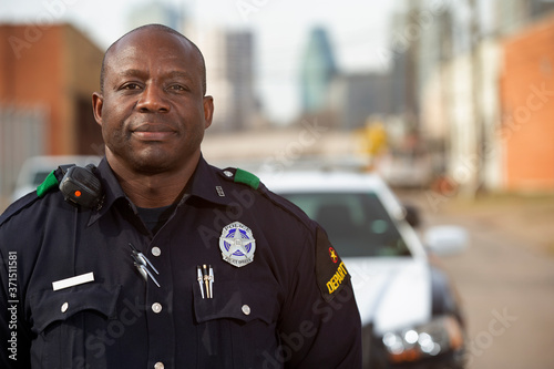 Portrait of Police officer Standing in street in front of squad car looking towards camera with arms behind his back 