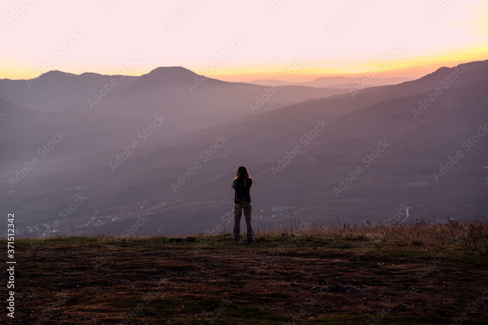 A young woman with a hiking backpack trekking poles stands high in the mountains and looks into the bluish valley. Sunset girl tourist stands back.