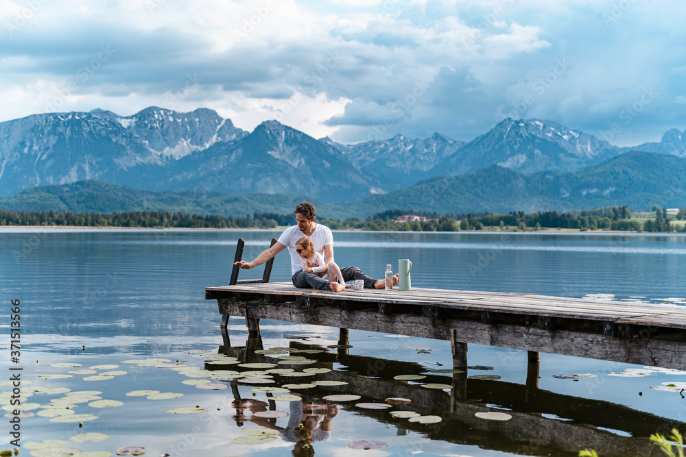 Father with daughter sitting on jetty over lake against mountains Father with daughter sitting on jetty over lake against mountains
