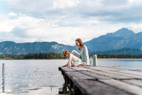 Mother with daughter sitting on jetty over lake against sky