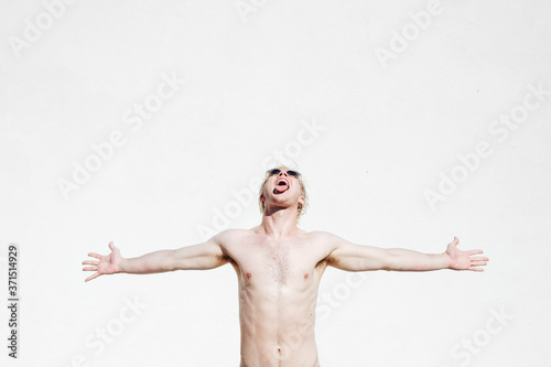 Shirtless young man with arms outstretched and mouth open standing against white wall