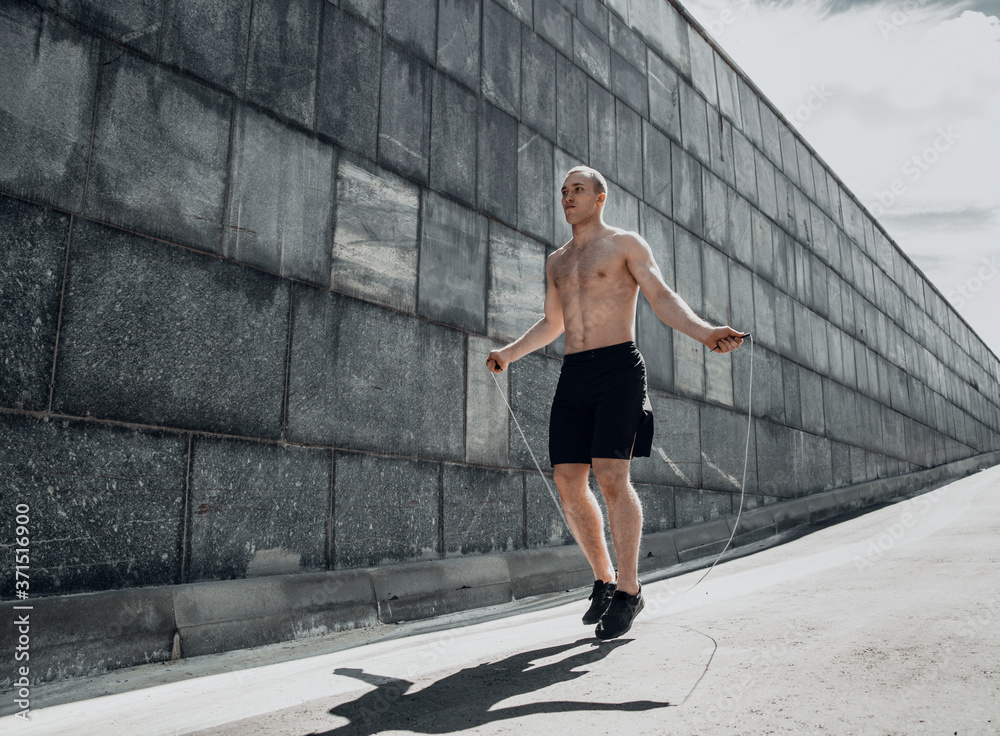 A young Caucasian guy with a naked torso jumps over a rope during a morning workout. Urban landscape.