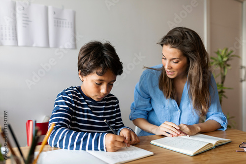 Female tutor assisting boy in writing homework on table at home photo