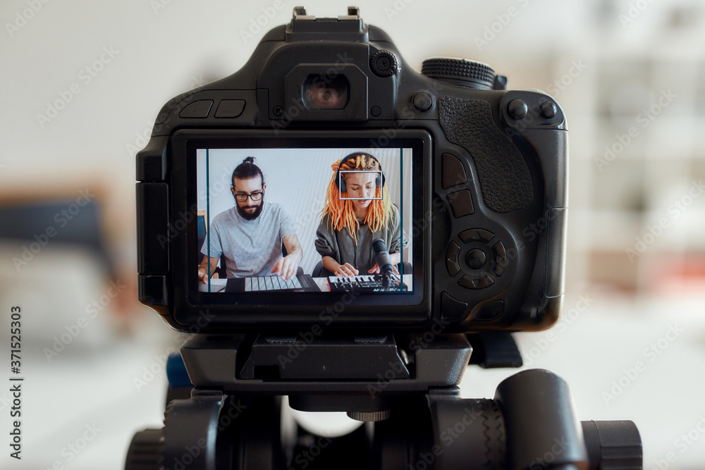 Woman with dreadlocks singing and playing. Female and male blogger recording making music using synthesizer, drum pad machine. Focus on camera screen