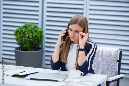 Beautiful young woman working on a laptop and talking on the phone smiling while sitting outdoors in a cafe. Young woman using laptop for work. Female freelancer working on a laptop in an outdoor cafe © Дмитрий Ткачук