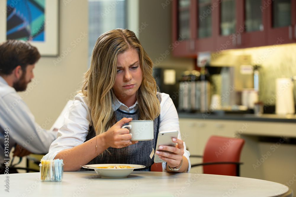 Young Caucasian woman looking at cellphone, while sitting in break area of office with lunch of soup and Hot Tea Young Caucasian woman looking at cellphone, while sitting in break area of office with lunch of soup and Hot Tea