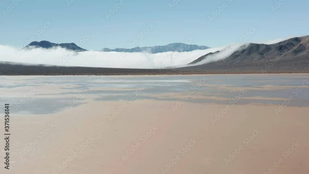 Sunrise on sunny winter day, 4K California. Aerial view of a drone flying over the flat wet cracked surface of Death Valley desert with high black mountains covered by thick fog clouds on background Sunrise on sunny winter day, 4K California. Aerial view of a drone flying over the flat wet cracked surface of Death Valley desert with high black mountains covered by thick fog clouds on background