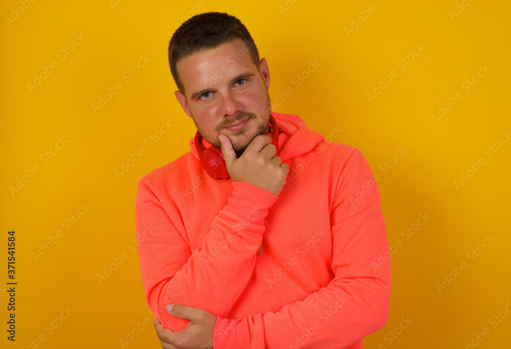 Portrait of thoughtful smiling man keeps hand under chin, looks directly at camera, listens something with interest, dressed casually, poses against gray wall. Youth concept. Portrait of thoughtful smiling man keeps hand under chin, looks directly at camera, listens something with interest, dressed casually, poses against gray wall. Youth concept.