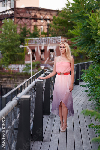 Beautiful blonde woman in pink strapless dress stands on walk way lined with greenery and abandoned building behind her