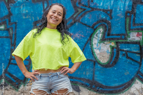 A young, happy girl in shorts and a bright T-shirt stands against the background of a wall with a blue graffiti drawing and smiles