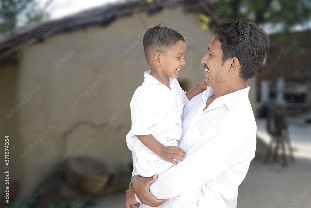 Happy rural indian young father and little son looking at each other and laughing wearing white traditional cloths with village background, copy space to write text.