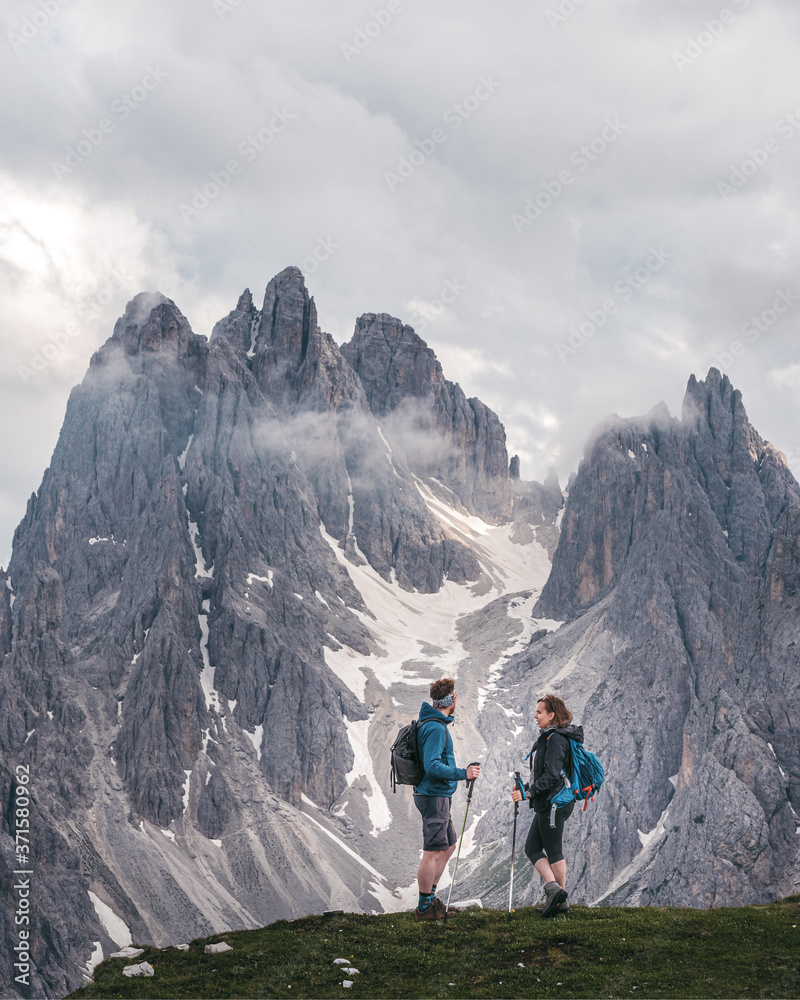 Two hikers standing and talking, with impressive rugged peaks of Cadini di Misurina mountain group towering on the background