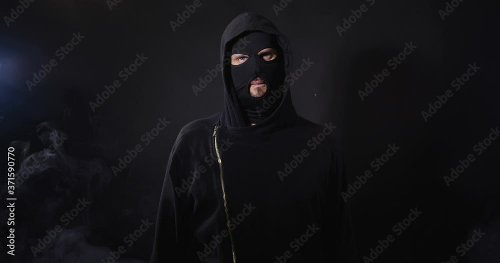 Portrait of robber in black clothes and mask looking aggressive to camera on black studio background with smoke. Security system concept. 4K 50 fps slow motion Portrait of robber in black clothes and mask looking aggressive to camera on black studio background with smoke. Security system concept. 4K 50 fps slow motion