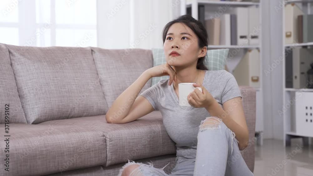 asian girl sitting by couch with hot tea is whiling away time at home. japanese girl looking into distance is having coffee and lost in thought. asian girl sitting by couch with hot tea is whiling away time at home. japanese girl looking into distance is having coffee and lost in thought.