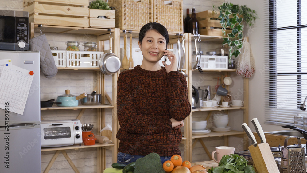 japanese housewife standing in kitchen holding cell phone is getting telephone sales call. asian woman taking a look at carrot while talking on phone is going to make lunch after she hangs up.