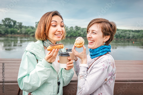 Two girl friends had a picnic in the city Park on the embankment. They eat eclairs and drink tea in take away boxes. Street food and communication concept