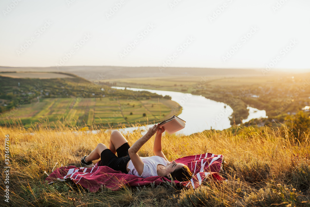 Young beautiful woman lying on a blanket on the grass on the hill and reading a book. She read a book with beautiful landscape on background. Copy space.