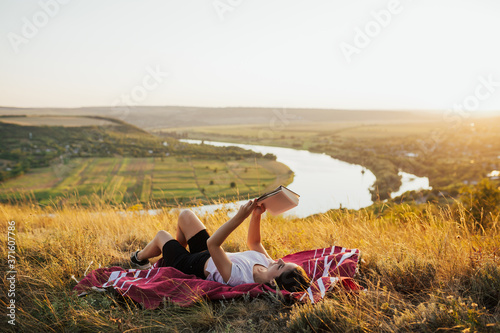 Obraz na płótnie Young beautiful woman lying on a blanket on the grass on the hill and reading a book. She read a book with beautiful landscape on background. Copy space. w salonie