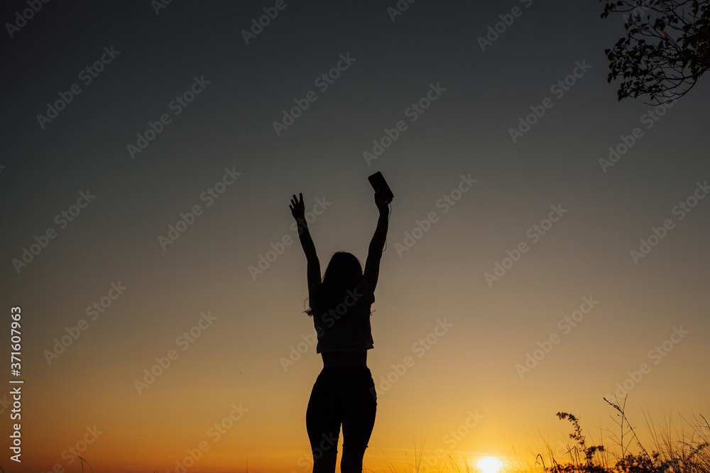 The girl at sunset. She stands back to the camera on the top of the mountain and looks at the sunset, welcomes the sun with her hands up. Girl listen the music.  Copy space.
