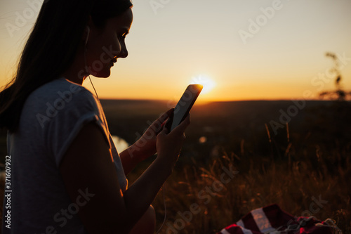 Attractive smiling young woman feeling happy while listening music in the headphone, during evening sunset. Close up. Copy space.