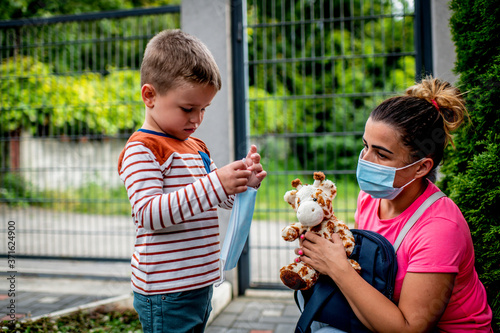 Caucasian Child getting ready to go to kindergarten in pandemic times, his mom is helping him to wear mask before he goes