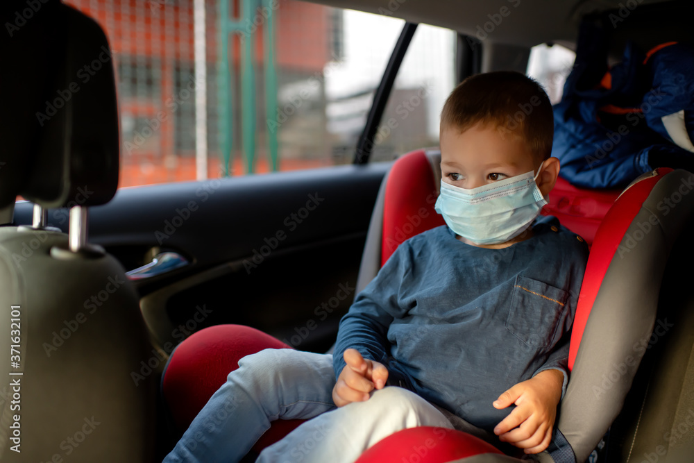 Cute little boy sitting at a car seat wearing protective mask