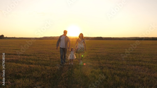 little daughter holds mom and dad by hands and jumps on meadow in park in rays of yellow sun. Family happiness. healthy baby playing with dad and mom on field in sunset light. Walking child in nature.