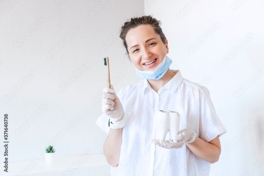 Smiling female dentist in white uniform holding white tooth and bamboo toothbrush, looking at camera against stomatology interior. Medicare. Tooth cure. Smiling female dentist in white uniform holding white tooth and bamboo toothbrush, looking at camera against stomatology interior. Medicare. Tooth cure.