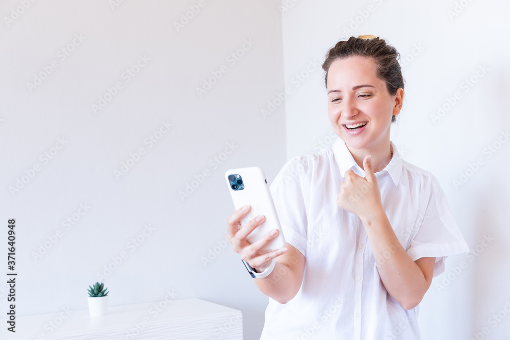 Young caucasian female in white blouse holding smartphone and showing thumbs up gesture on home interior. New technology. Modern lifestyle. Space for text. Young caucasian female in white blouse holding smartphone and showing thumbs up gesture on home interior. New technology. Modern lifestyle. Space for text.