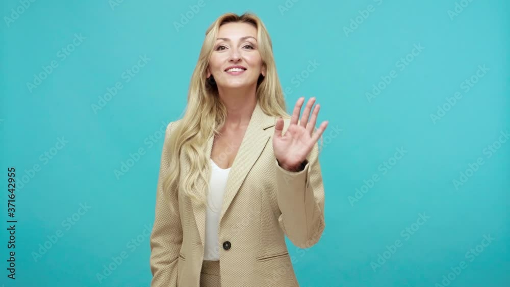 Friendly positive middle aged blond woman in stylish suit waving hand looking at camera with toothy smile, saying hi, greeting. Indoor studio shot isolated on blue background Friendly positive middle aged blond woman in stylish suit waving hand looking at camera with toothy smile, saying hi, greeting. Indoor studio shot isolated on blue background