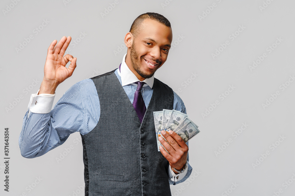 Portrait of excited young african american man holding cash money and showing OK sign over light grey background Portrait of excited young african american man holding cash money and showing OK sign over light grey background