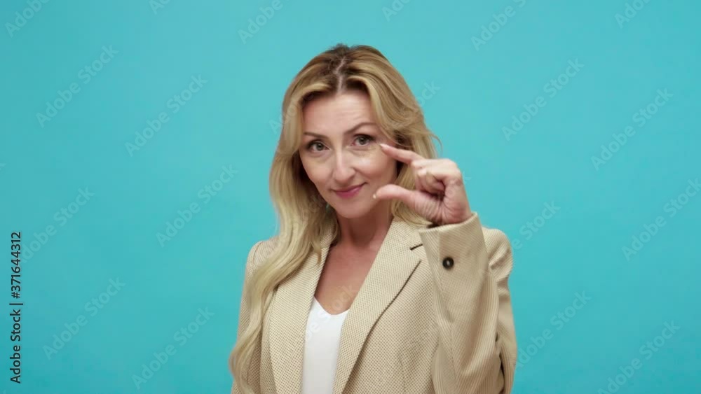 Closeup blond middle aged woman in jacket showing a little bit gesture looking at camera with smile, showing minimum sign, low rating. Indoor studio shot isolated on blue background Closeup blond middle aged woman in jacket showing a little bit gesture looking at camera with smile, showing minimum sign, low rating. Indoor studio shot isolated on blue background