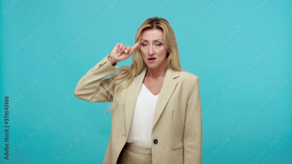 I am stupid. Tired depressed adult woman in business style suit twisting finger near temple, making stupid gesture and sadly looking at camera. Indoor studio shot isolated on blue background I am stupid. Tired depressed adult woman in business style suit twisting finger near temple, making stupid gesture and sadly looking at camera. Indoor studio shot isolated on blue background