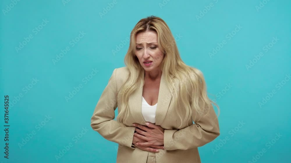 Adult blond woman in business style jacket grimacing and writhing in pain feeling stomach ache, indigestion, sore periods, miscarriage. Indoor studio shot isolated on blue background Adult blond woman in business style jacket grimacing and writhing in pain feeling stomach ache, indigestion, sore periods, miscarriage. Indoor studio shot isolated on blue background