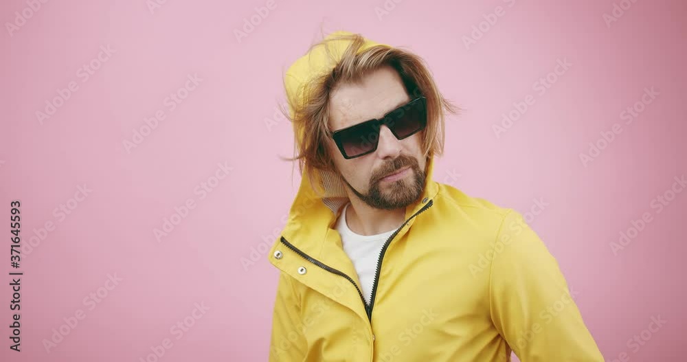 Serious bearded man in trendy sunglasses and yellow raincoat posing on camera in studio. Mature male standing over pink background with hood on head. Serious bearded man in trendy sunglasses and yellow raincoat posing on camera in studio. Mature male standing over pink background with hood on head.
