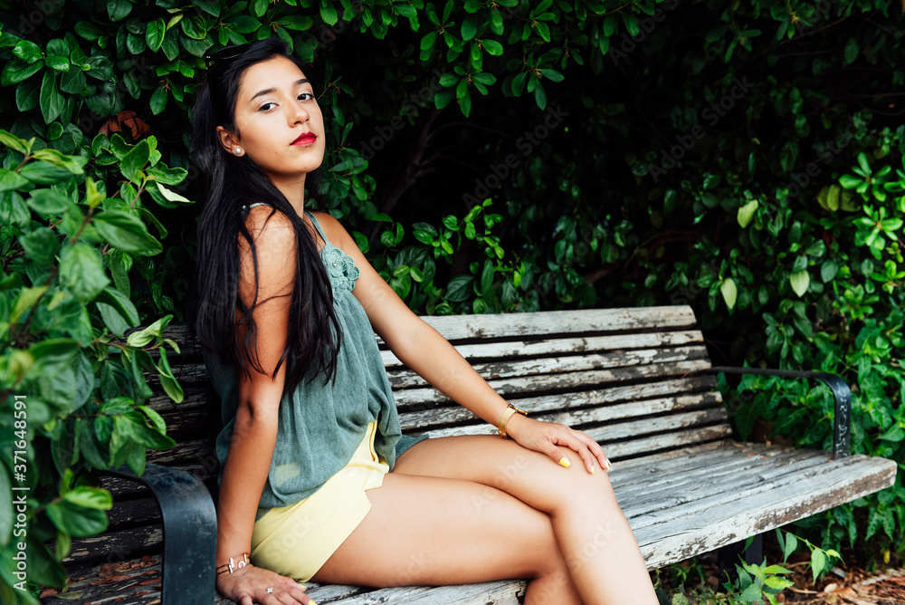 Latin young girl with long black hair and blue dress, sitting on wooden bench among greenery, looking at camera Latin young girl with long black hair and blue dress, sitting on wooden bench among greenery, looking at camera