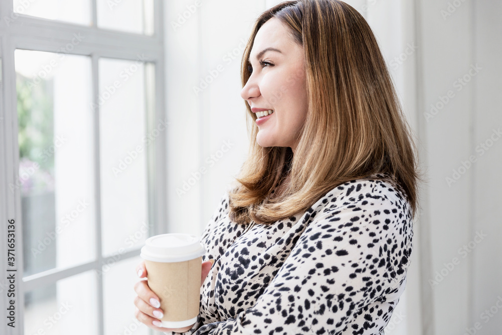 Smiling Asian business woman stands with a craft glass of coffee and looks out the window. Smiling Asian business woman stands with a craft glass of coffee and looks out the window.