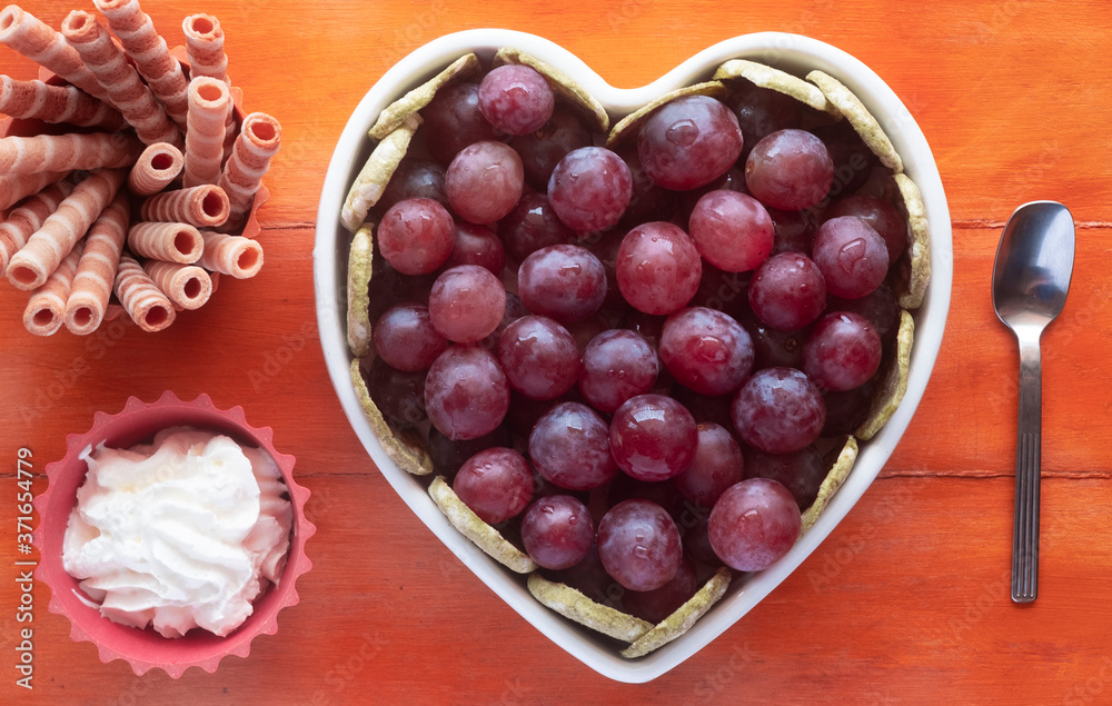 Close-up view of the white heart-shaped plate filled with ripe red grapes, accompanied by cream and biscuits. Wooden background Close-up view of the white heart-shaped plate filled with ripe red grapes, accompanied by cream and biscuits. Wooden background