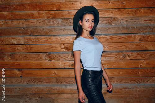 Profile portrait of a fashion girl in hat, her hair straight dressed in leather pants and t shirt near the wooden wall, walking at night.