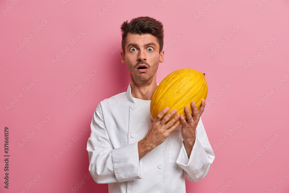 Stunned emotional male cook holds delicious sweet ripe melon, going to prepare tasty dessert, stares at camera with shock, wears uniform, isolated on pink background. Healthy eco food, organic farming